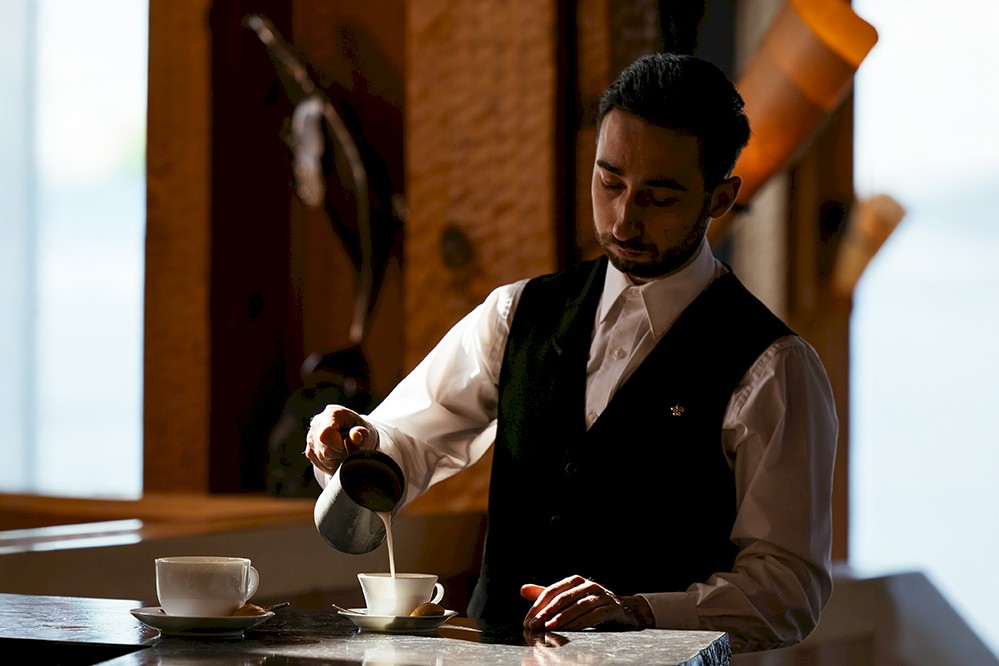 A person is pouring a hot beverage into a cup on a marble counter in a restaurant setting. Warm lighting and wooden decor create a comfortable atmosphere.