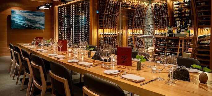 A long, elegant dining table set with wine glasses, plates, and menus is surrounded by chairs. The room features wooden walls lined with wine racks filled with bottles. A framed picture of an ocean scene hangs on the wall.