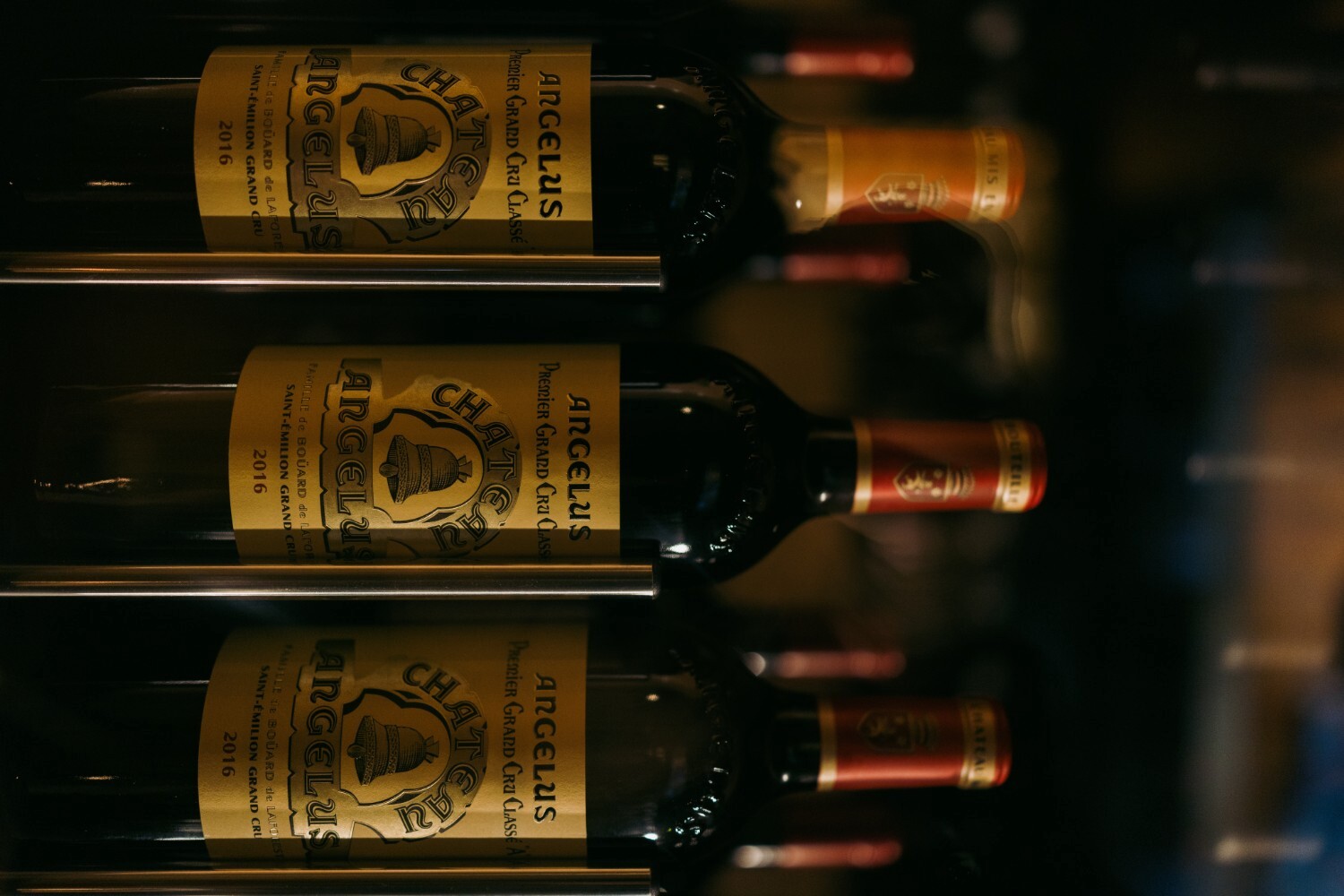 Three bottles of Château Angélus wine are displayed on a rack. The labels feature a bell graphic and gold accents.