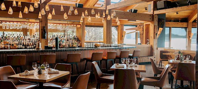 A cozy, upscale bar interior featuring wooden beams and warm lighting. The seating area includes neatly arranged tables with leather chairs, set with wine glasses and cloth napkins. The bar area is well-stocked, and modern pendant lights hang from the ceiling. Large windows provide ample natural light.
