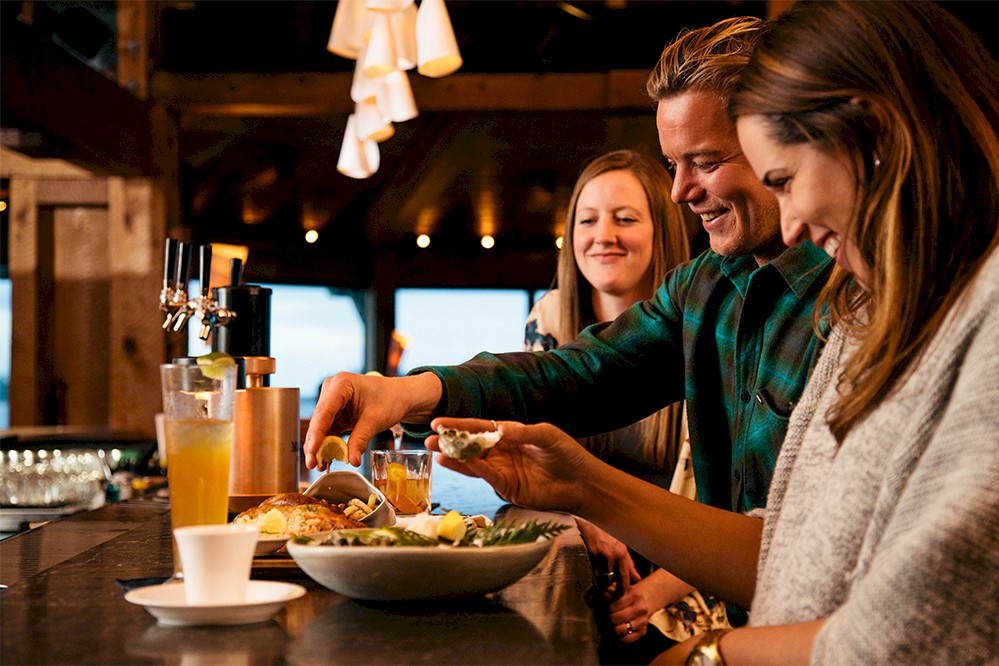 Three individuals are seated at a bar with drinks and food. One is squeezing lemon over a dish. The setting appears warm and cozy, with hanging lights and wooden decor.