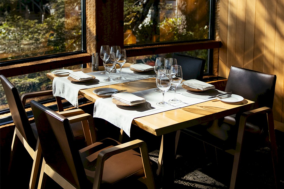 A sunlit dining table set for four with neatly arranged tableware and wine glasses. The table features a white cloth runner and overlooks a window showcasing a view of lush greenery.