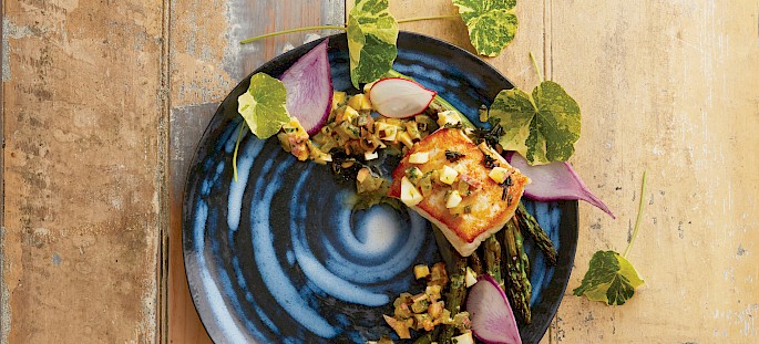 A beautifully plated dish featuring grilled halibut on a blue spiral plate, accompanied by grilled asparagus, radish slices, and edible flowers on a rustic wooden table.