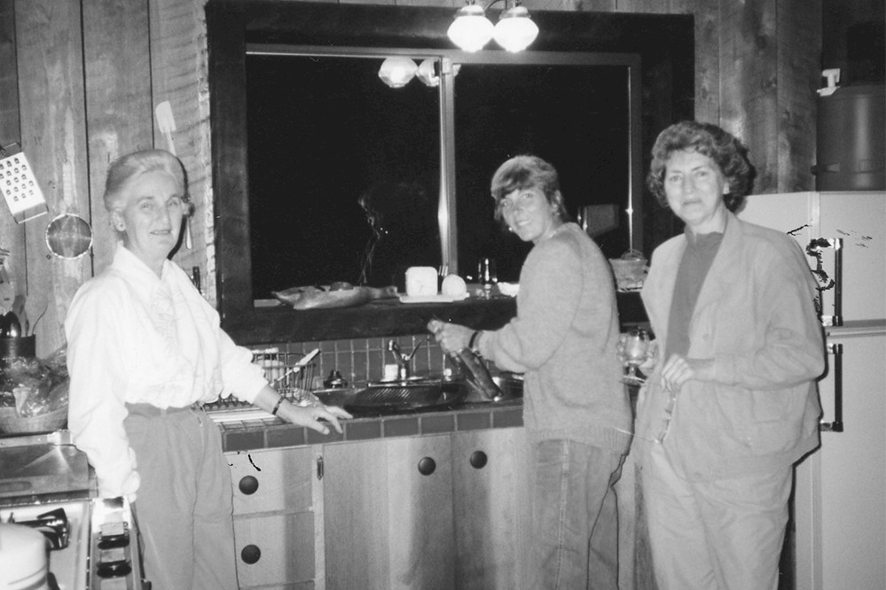 A group of three people are gathered in a rustic kitchen. One is at the sink with a faucet, and the others stand nearby. The kitchen features wooden walls and a large window. The atmosphere is casual and relaxed.