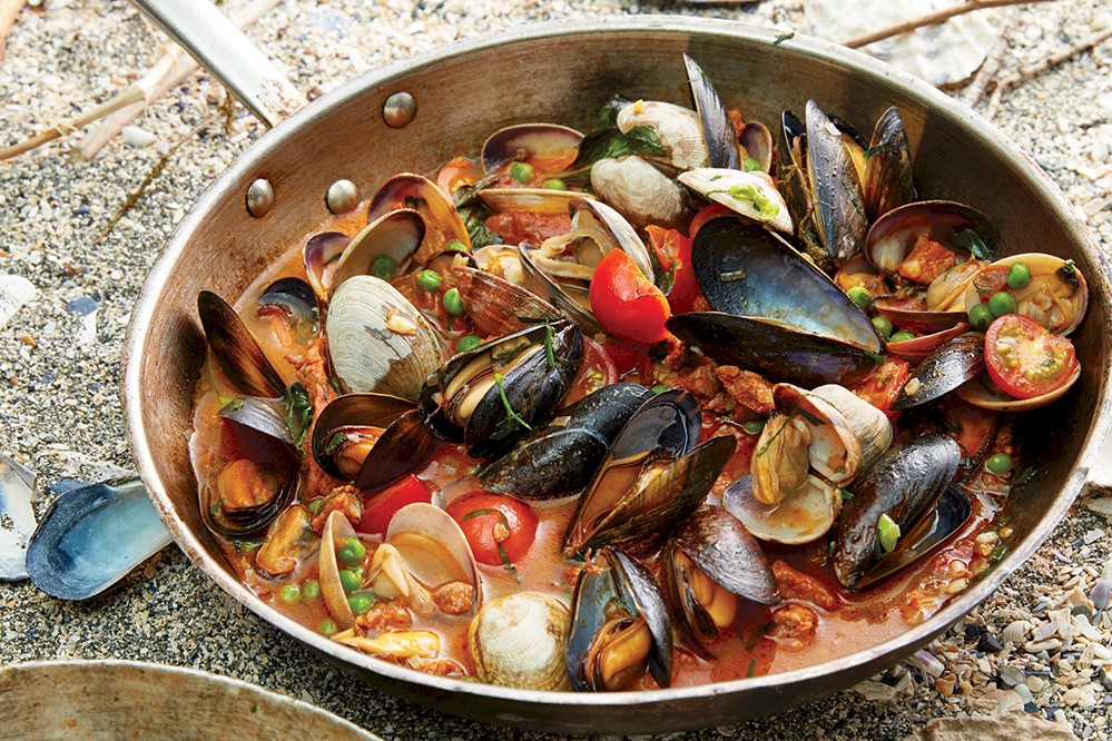 A pan filled with a seafood stew, featuring mussels, clams, cherry tomatoes, and peas in a red broth. The pan rests on the beach with sand and shells scattered around it.