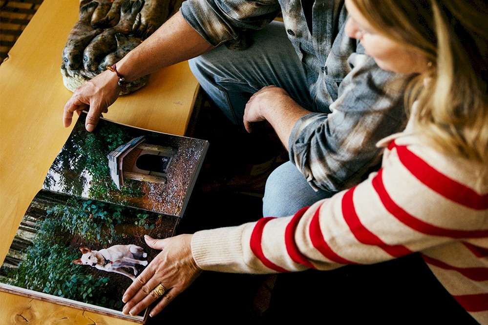 Two people sit at a table, flipping through the Wickaninnish Cookbook. The visible page shows a dog near a wooden dog house in a lush, green setting.