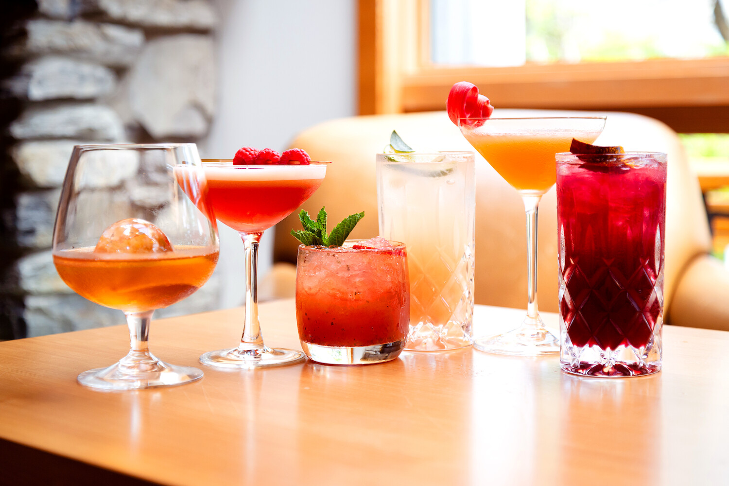 A selection of five colorful cocktails displayed on a wooden table, including drinks with garnishes like raspberries, a mint sprig, and citrus slices. The background shows a blurred view of a stone wall and a window with natural light.
