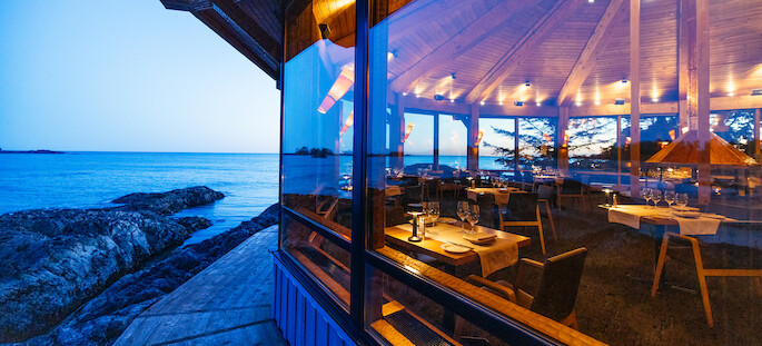 A cozy restaurant with large windows offers a view of the ocean and rocky shoreline at dusk. Inside, tables are set with white tablecloths, wine glasses, and candles, creating a warm and inviting ambiance.